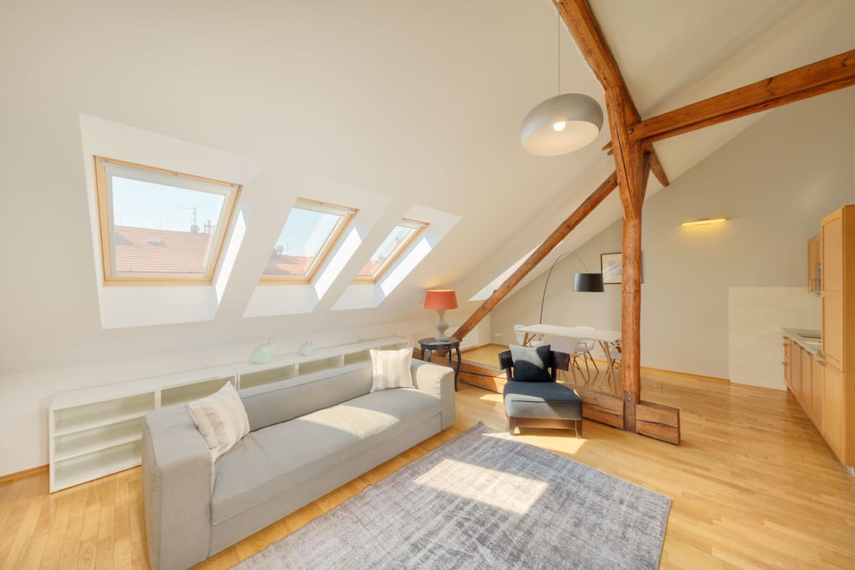 Eine moderne Loftlounge mit schrägem Dach, Dachfenstern, einem grauen Sofa, Holzboden und einer kleinen Essecke.