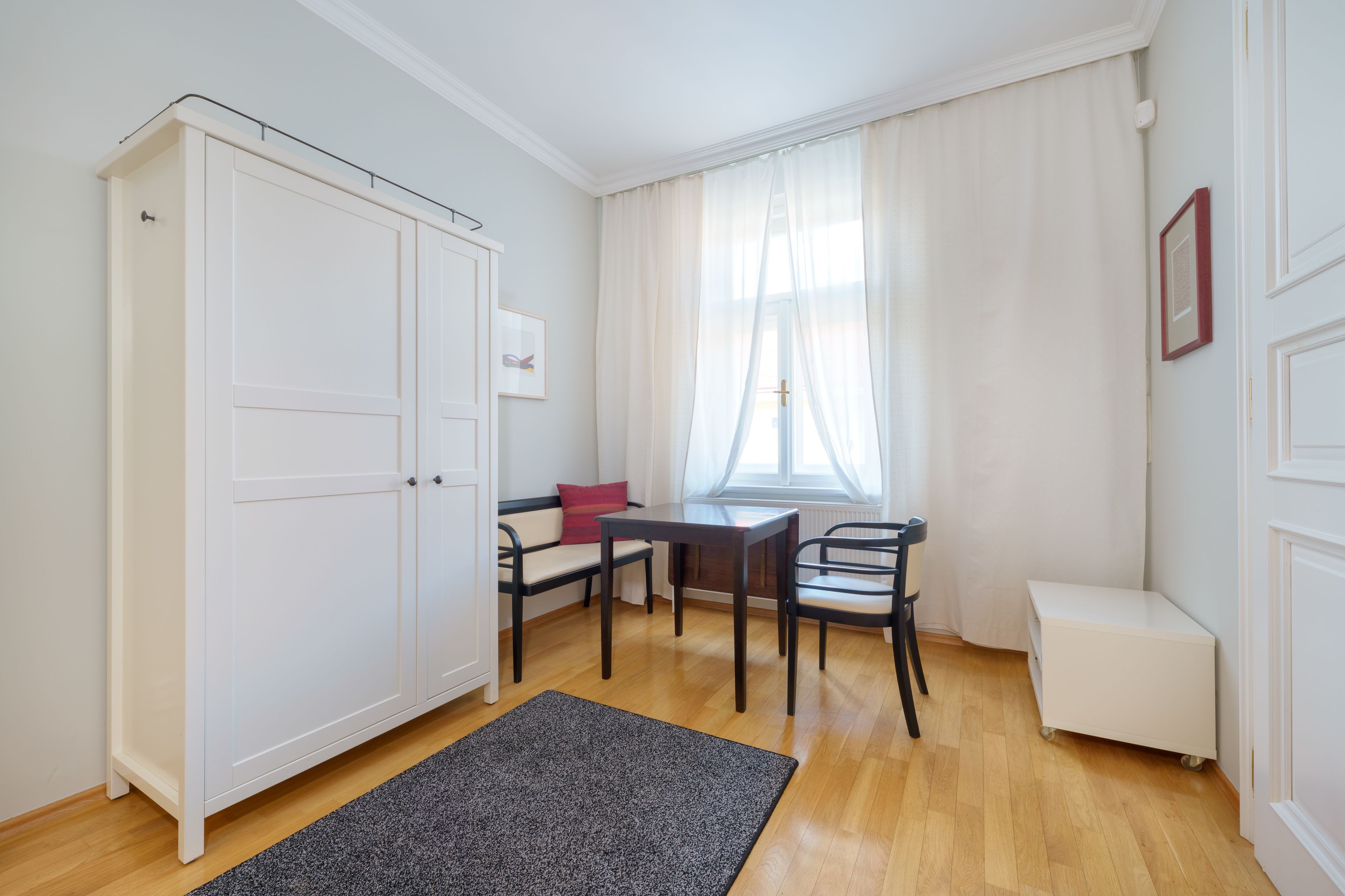 A room with a large white wardrobe, a small desk with a chair, a bench, and a window with sheer curtains.