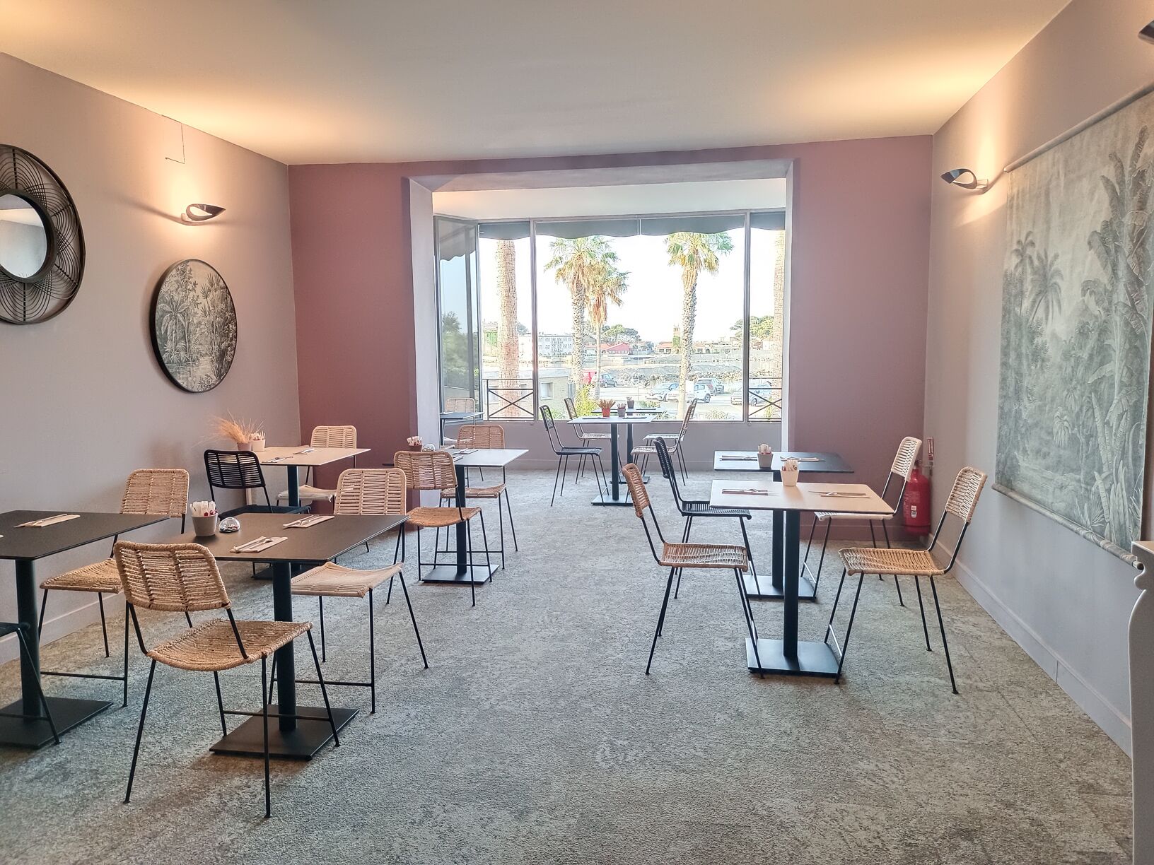 A modern dining area with multiple tables and chairs, large windows offering a view of palm trees and buildings, and artwork on the walls.
