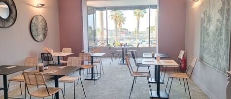 A modern dining area with multiple tables and chairs, large windows offering a view of palm trees and buildings, and artwork on the walls.