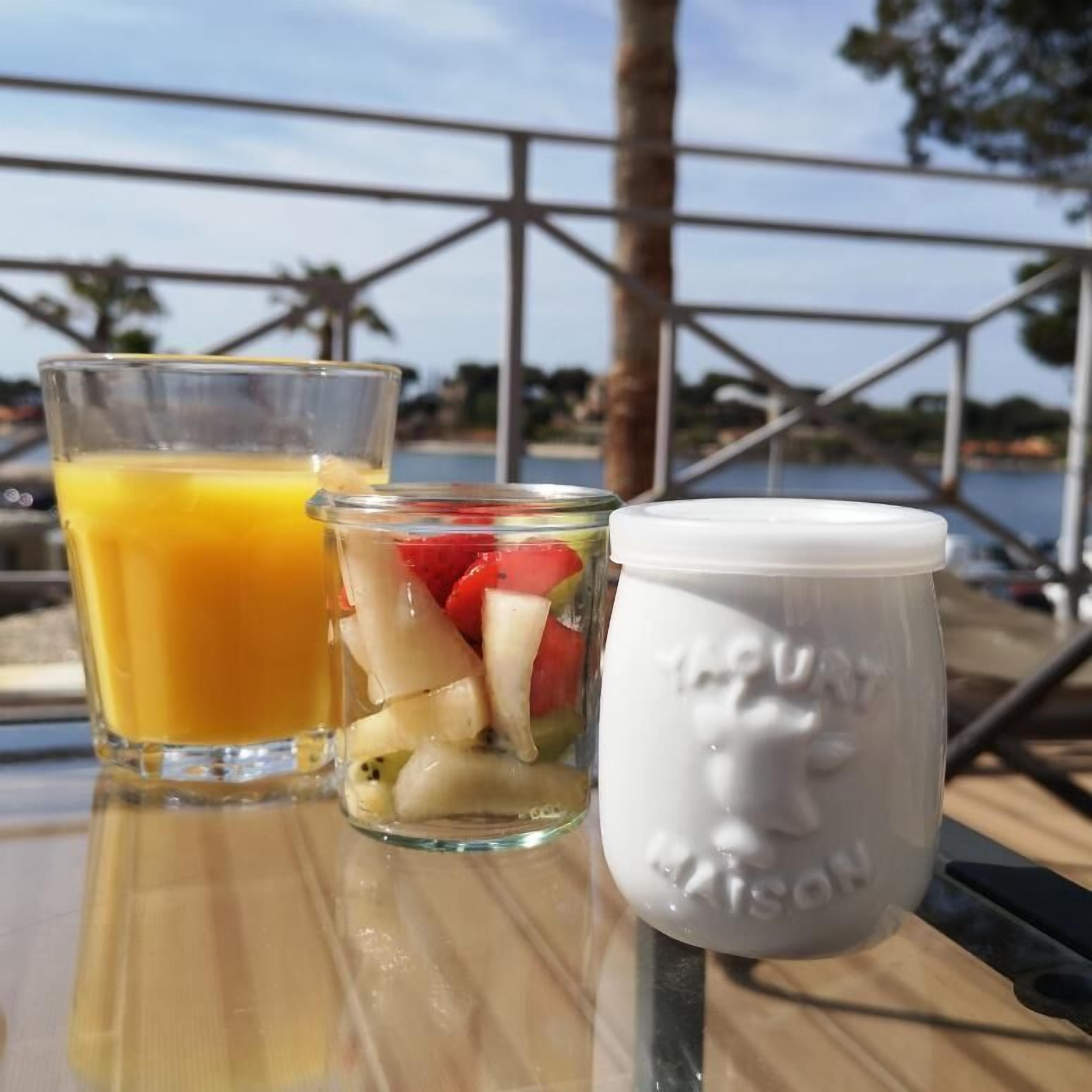 Una mesa con un vaso de jugo de naranja, un tarro de ensalada de frutas variadas y una taza blanca con un diseño de rostro.