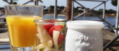 Una mesa con un vaso de jugo de naranja, un tarro de ensalada de frutas variadas y una taza blanca con un diseño de rostro.