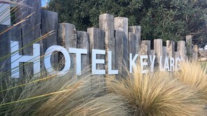 A wooden fence with the text "HOTEL KEY LARGO" partially obscured by tall grasses and rocks. - Key Largo (Bandol)