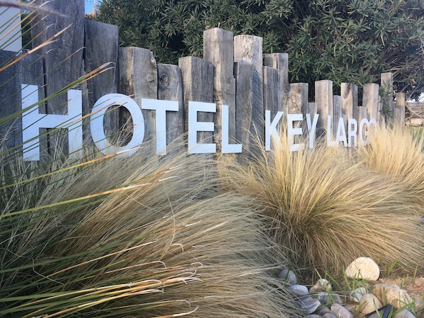 A wooden fence with the text "HOTEL KEY LARGO" partially obscured by tall grasses and rocks.