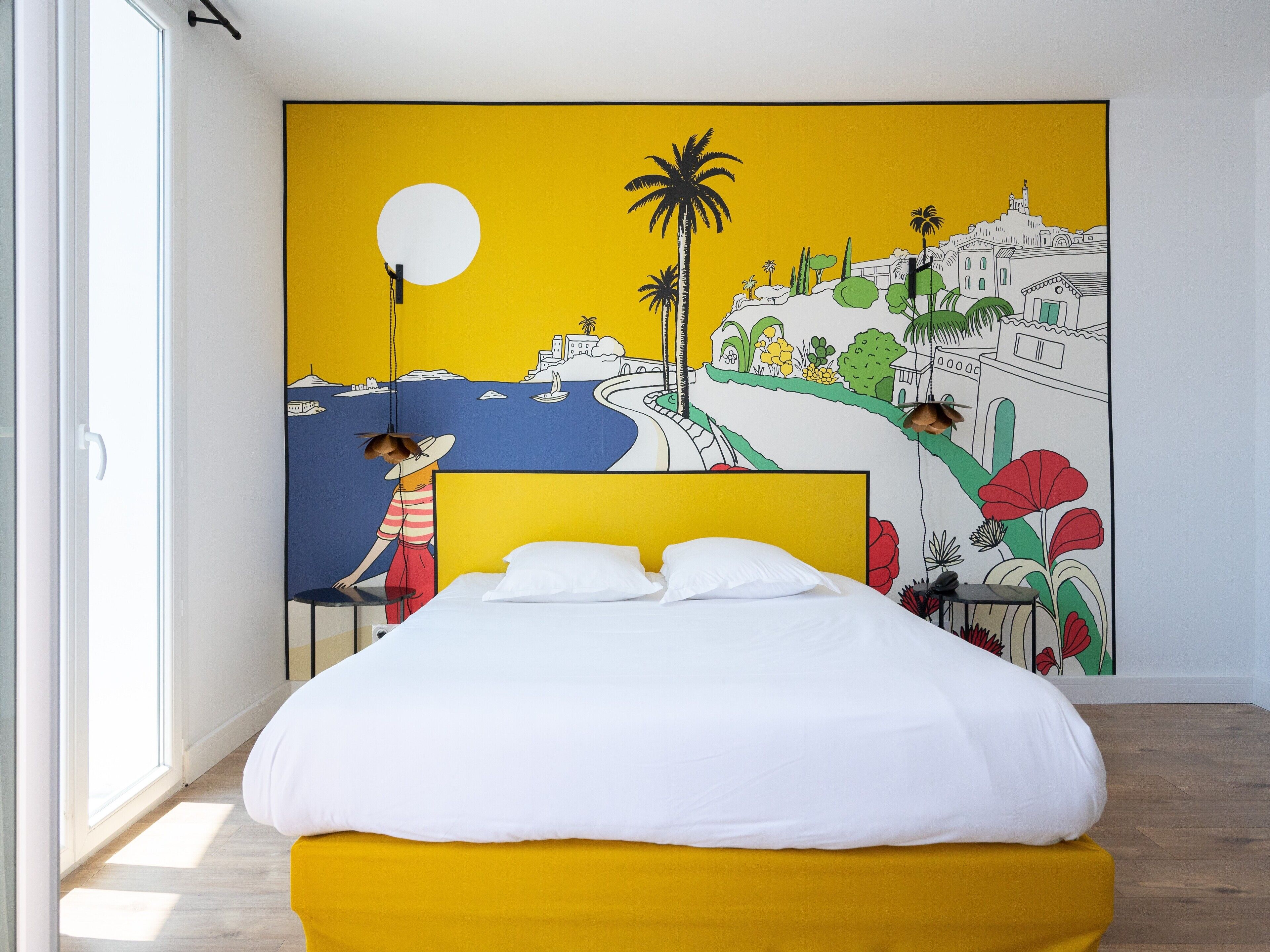Une chambre à coucher avec un lit, une tête de lit jaune et une grande fresque murale représentant un paysage côtier avec des palmiers et des bâtiments.