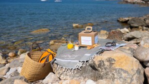A beach setting with a wicker basket, a towel, a bottle of orange juice, a newspaper, and a small box on a rock.