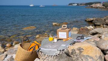 Une plage, avec un panier en osier, une serviette, une bouteille de jus d’orange, un journal et une petite boîte posée sur un rocher.
