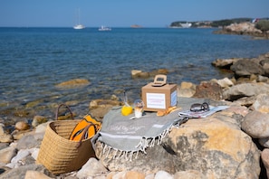 A beach setting with a wicker basket, a towel, a bottle of orange juice, a newspaper, and a small box on a rock.