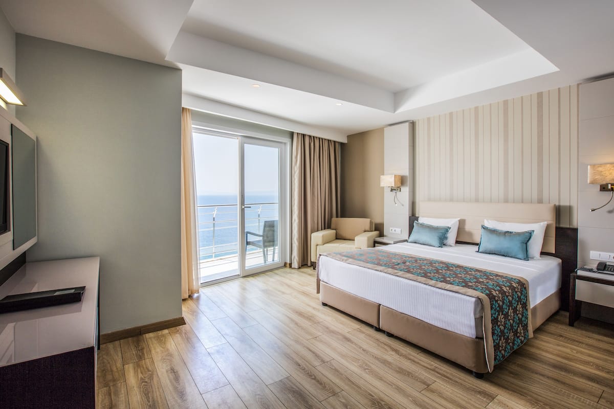 A hotel room with a large bed, a desk, a chair, and a balcony with a view of the sea.