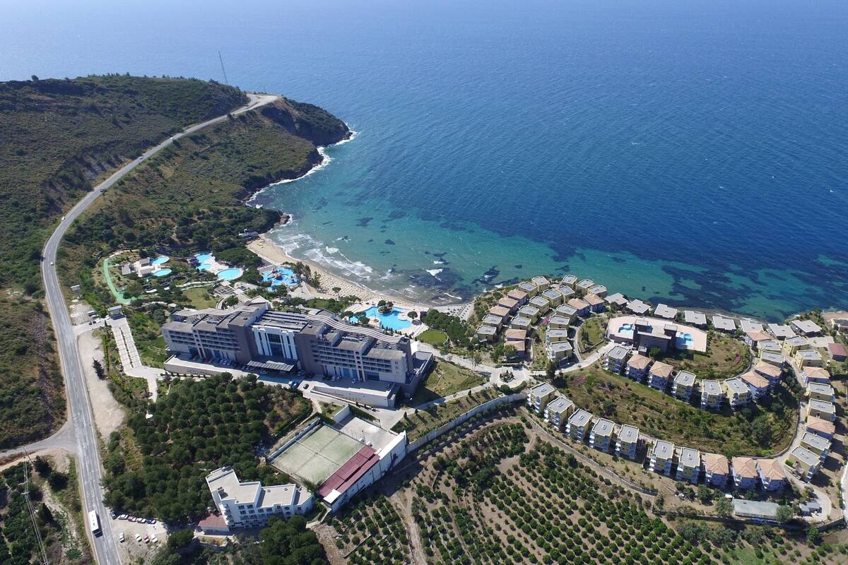 An aerial view of a coastal resort with a large building, swimming pools, and a residential area with villas.
