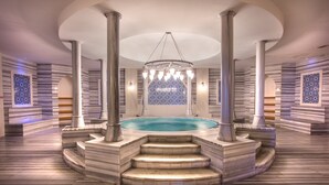 Une piscine intérieure luxueuse, avec des marches et des colonnes en marbre, entourée de murs décorés et surmontée d’un lustre central.