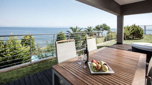 Une table en bois, sur laquelle se trouvent une assiette de fruits et des verres à vin, est placée sur un balcon donnant sur la mer.