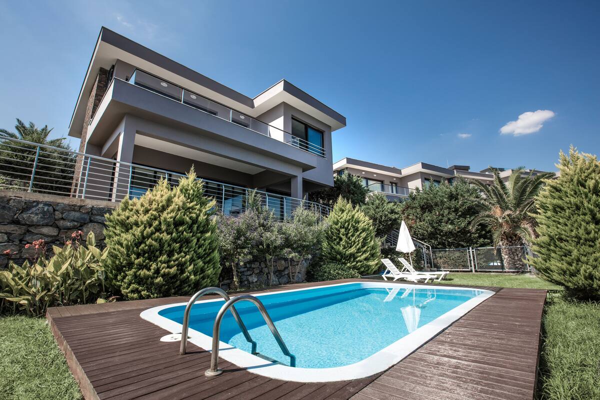 A modern two-story house with a swimming pool, surrounded by greenery and a stone wall.