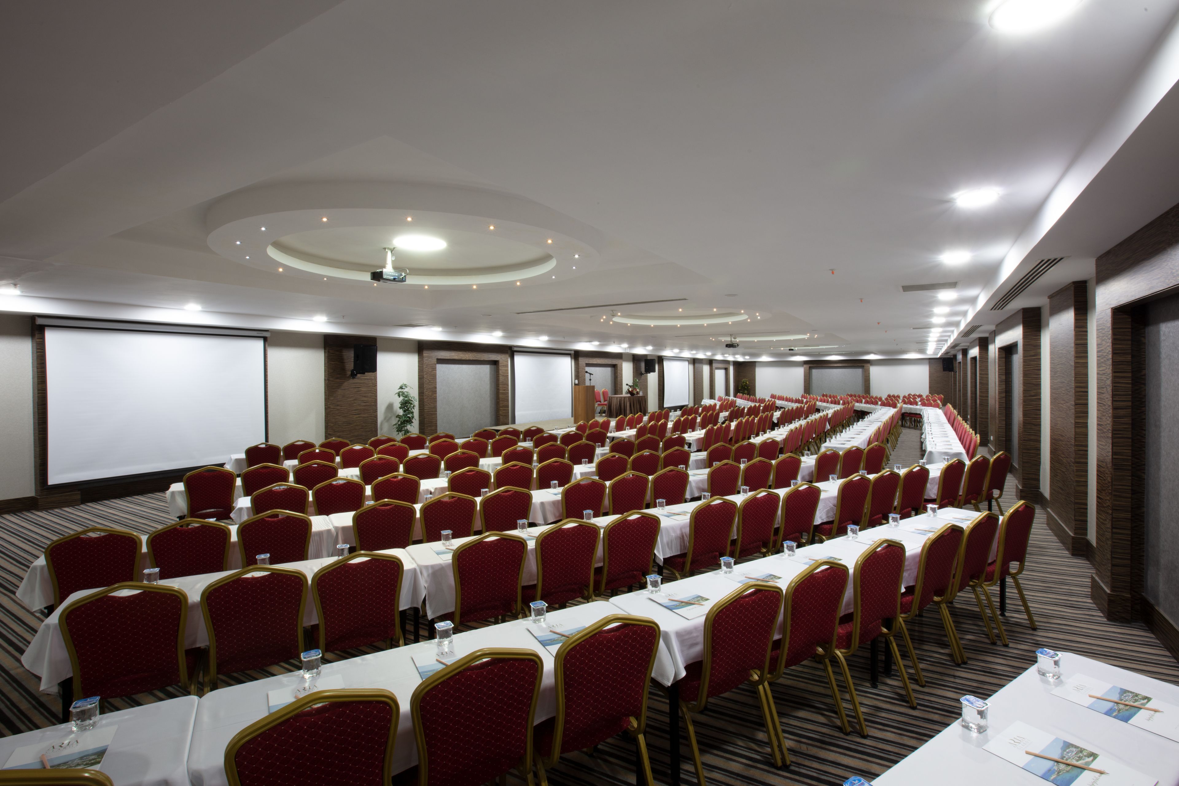 Een ruime congreszaal met rijen tafels en stoelen, een projectiescherm en een trap naar een hoger niveau.