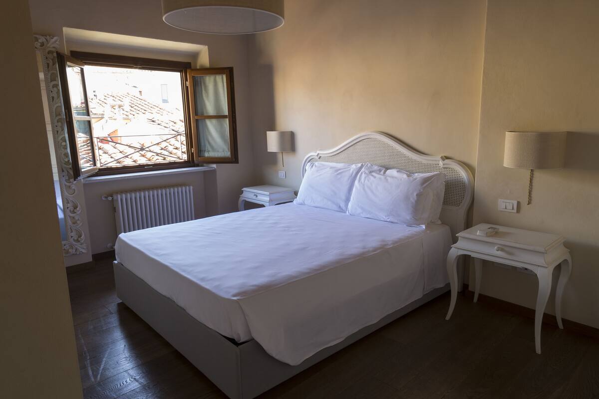 A neatly made bed in a hotel room with a window, a bedside table with a lamp, and a radiator.