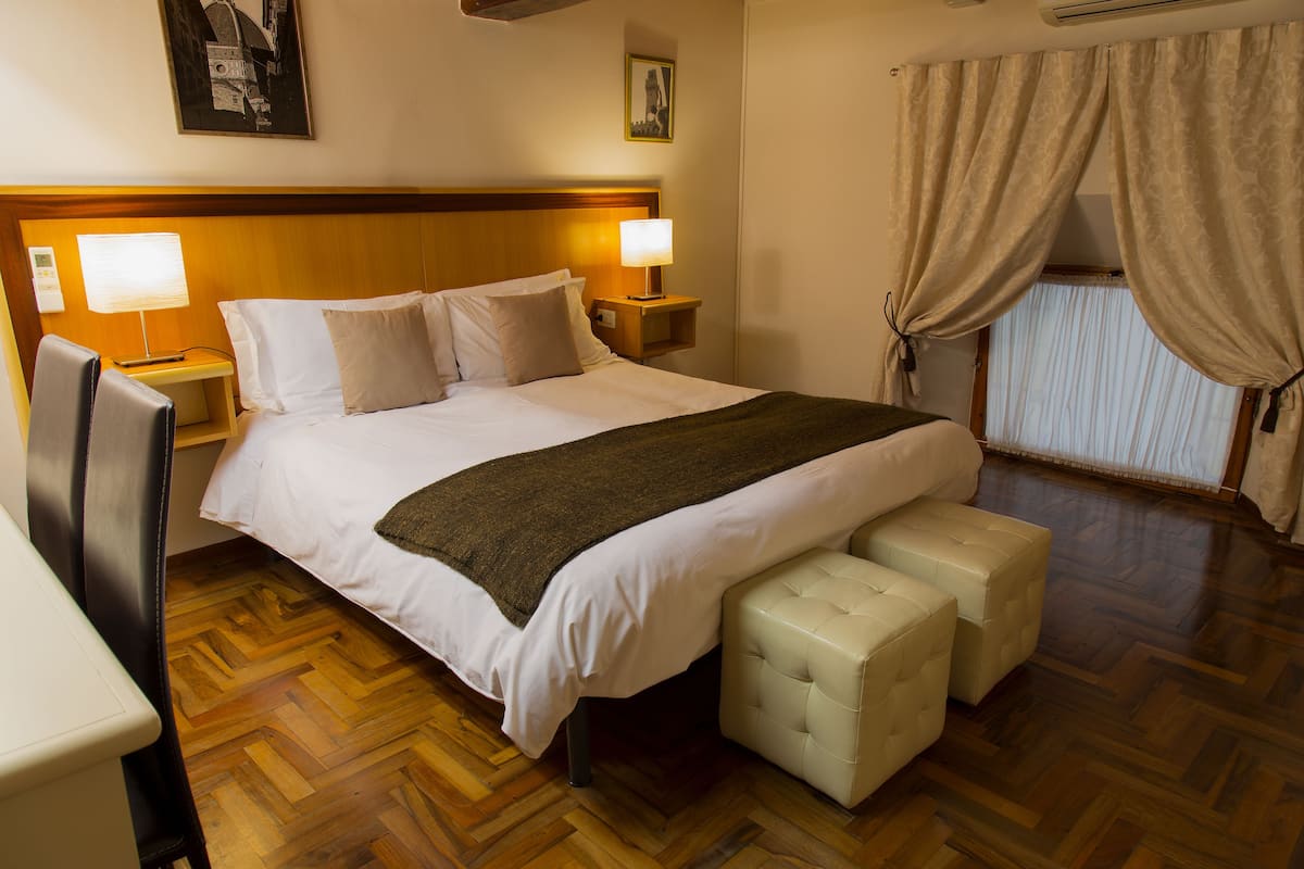 A hotel room with a large bed, two bedside lamps, a wooden headboard, a small bedside table, a chair, a window with curtains, and a wooden parquet floor.
