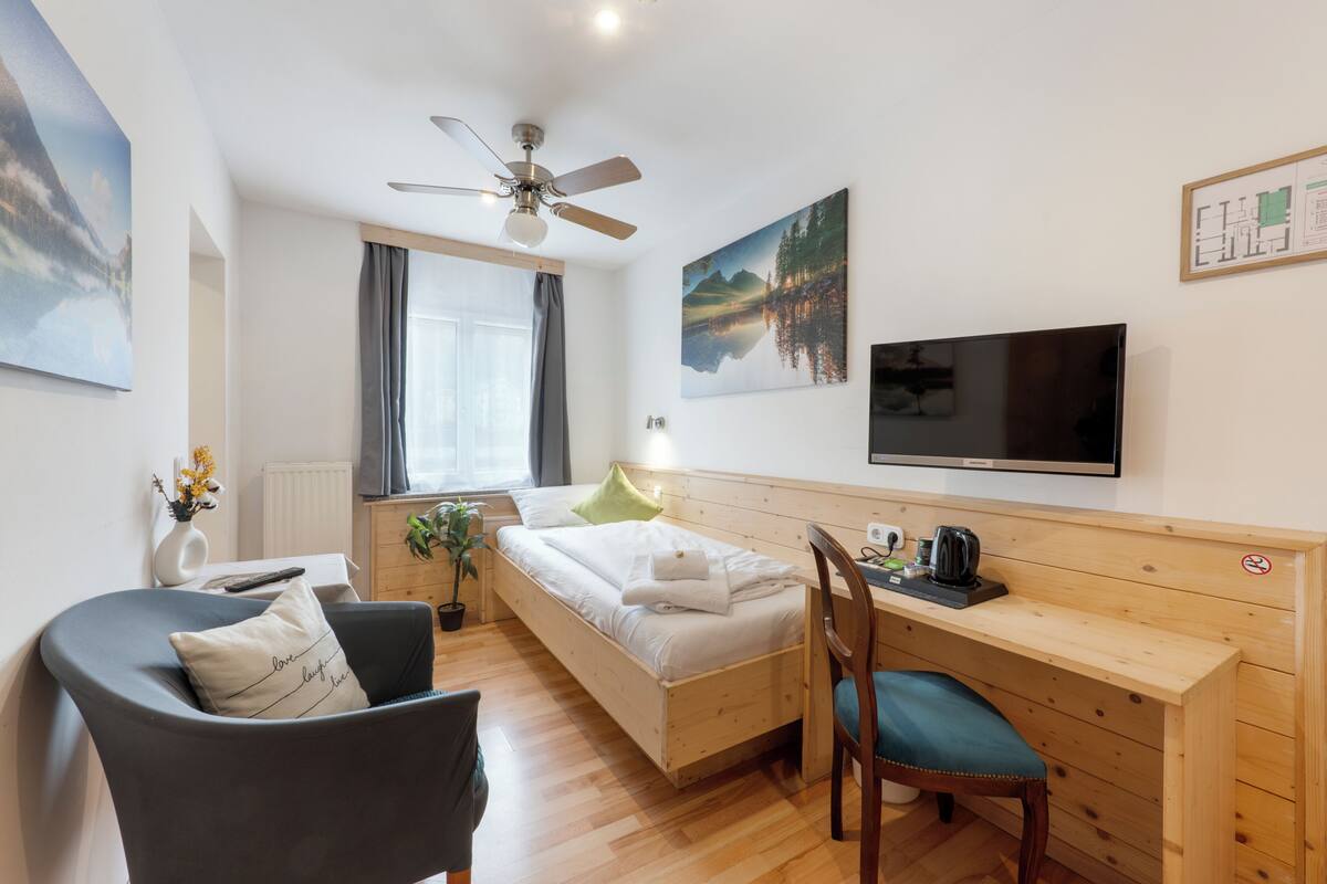 A room with a bed, a desk, a chair, a TV, a ceiling fan, and a window with curtains.