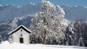 Een klein, besneeuwd kapelletje met een kruis aan de voorkant, omringd door sneeuwzware bomen en bergen op de achtergrond.