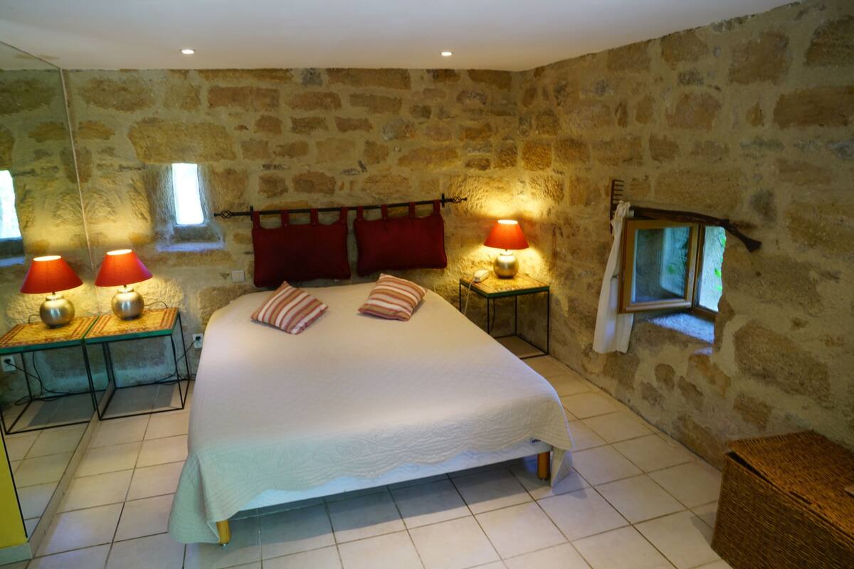 Une chambre avec un mur en pierre, un lit avec des oreillers, deux tables de chevet avec des lampes et une fenêtre où est accrochée une serviette.
