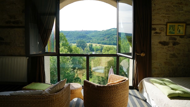 Een kamer met een groot rondboogvenster met uitzicht op een groen landschap. Er staat een bed met een groene deken en een rieten stoel met kussens.