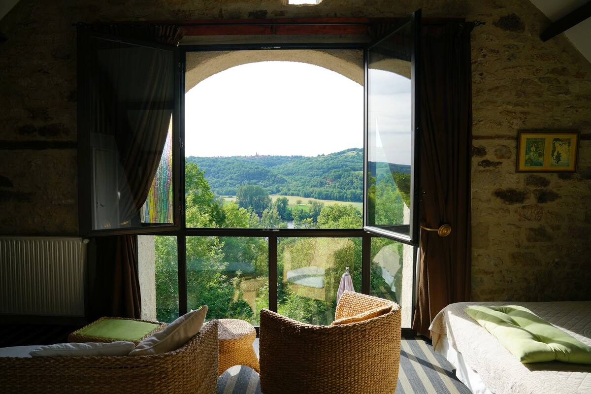 Une pièce dotée d’une grande fenêtre en arc d’où l’on peut admirer un paysage verdoyant. On y trouve un lit recouvert d’une couverture verte et une chaise en osier agrémentée de coussins.