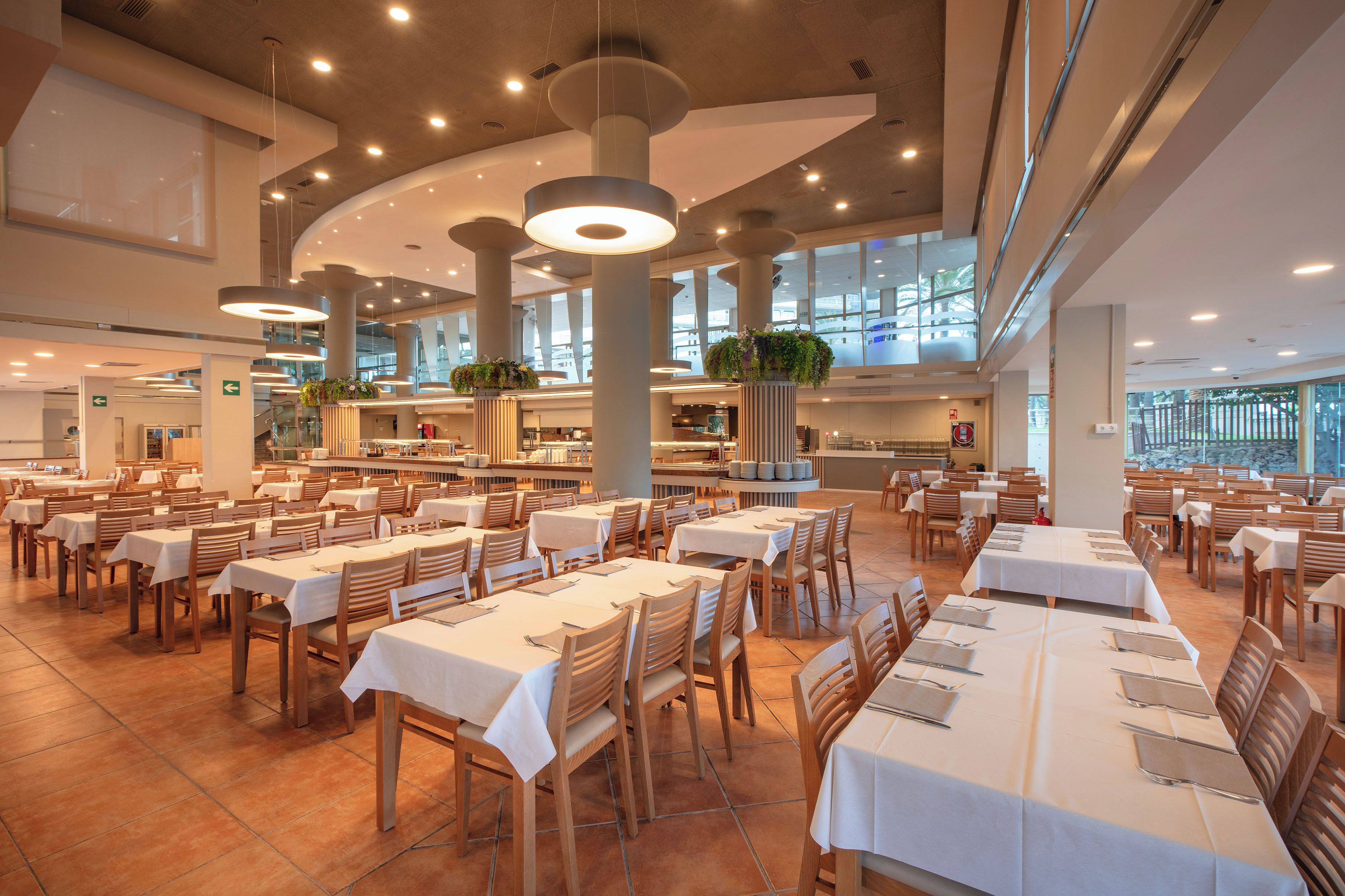 A spacious dining area with multiple tables and chairs, large windows, and hanging light fixtures.