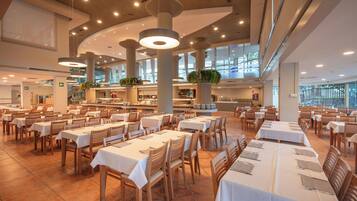A spacious dining area with multiple tables and chairs, large windows, and hanging light fixtures.