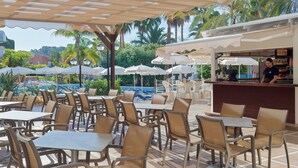 An outdoor café with tables and chairs, a pool in the background, and a bar area. - Ponient Dorada Palace by PortAventura World (Salou)