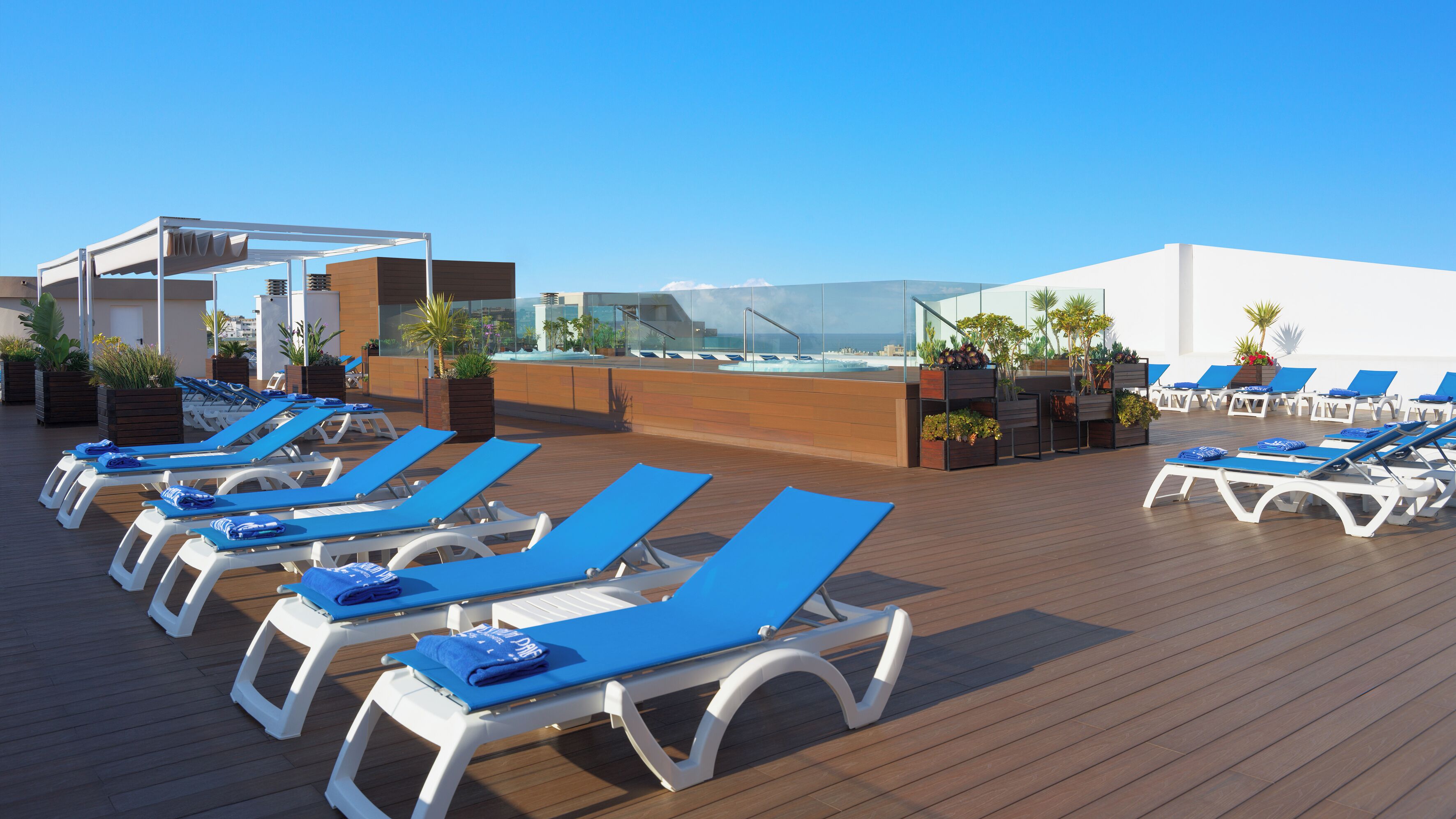 Un área de lounge en la azotea con sillones de sol y un bar con paredes de vidrio.