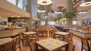 Une cafétéria spacieuse, dotée de nombreuses tables et chaises, de grands luminaires suspendus et d’un comptoir central où un chef prépare les plats.