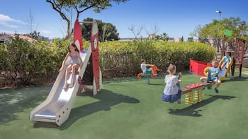 Children can play on the slide and seesaw in this well-maintained outdoor play area surrounded by trees and greenery.