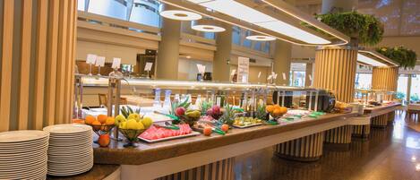 A buffet with various fruits and vegetables, a counter with plates, and a modern interior with large windows and columns.