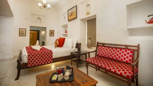 A hotel room with a bed, a wooden bench, a small table with a tray, and a chandelier. - Amritara Chandra Mahal Haveli (Nadbai)