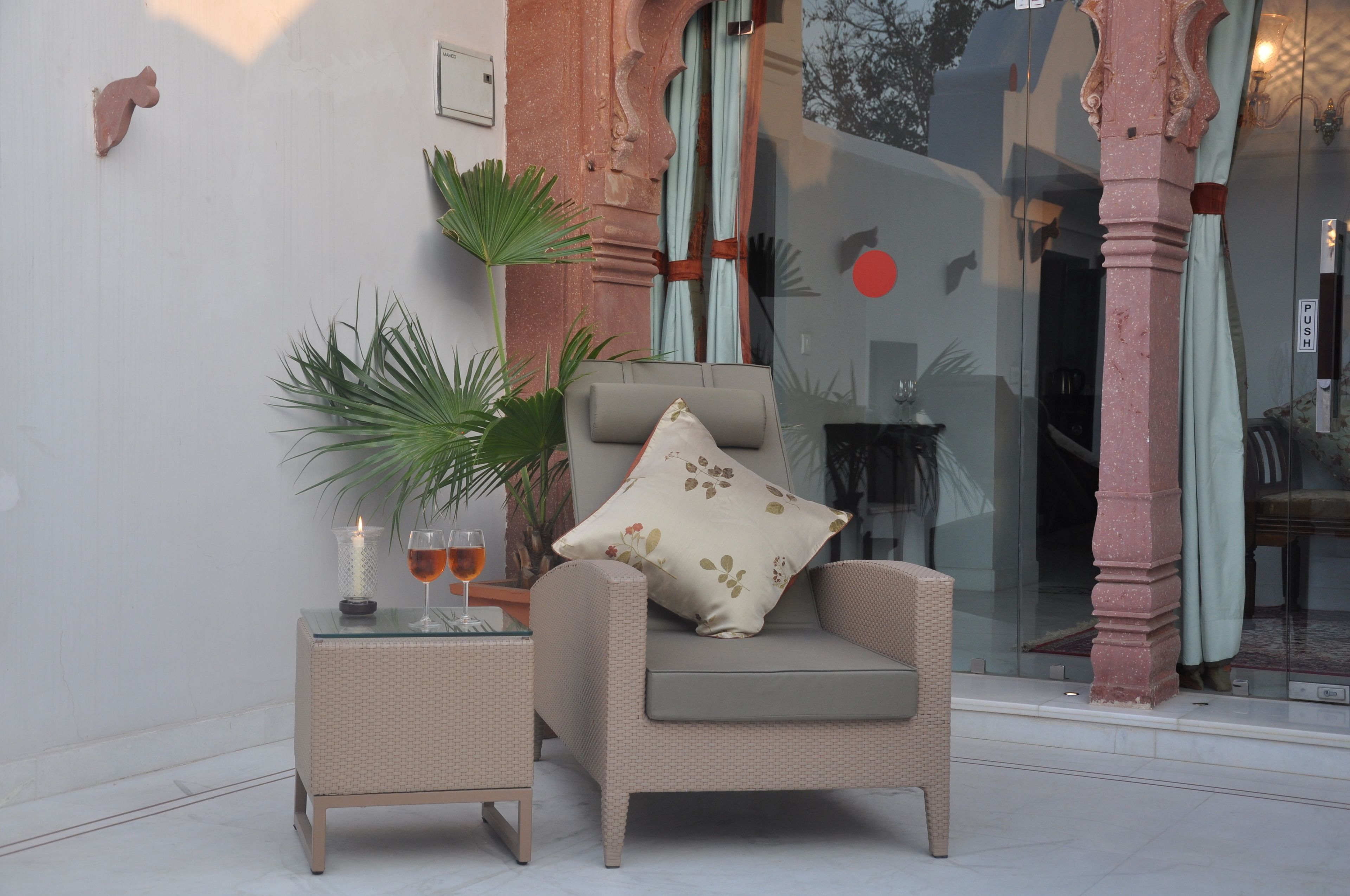 A wicker armchair with a cushion, a glass table with a candle and a glass, and a potted plant.
