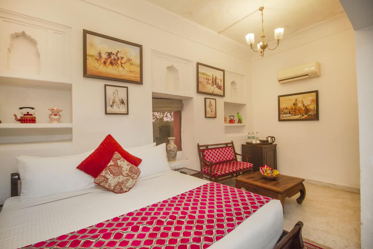 Quarto de hotel com uma cama grande, uma mesa de centro de madeira, um edredom com padrão vermelho e branco, e várias obras de arte emolduradas nas paredes.