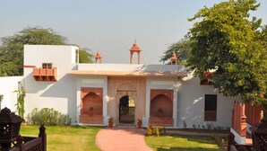 A traditional building with a central archway, a pathway leading to the entrance, and a surrounding garden with potted plants and a tree. - Amritara Chandra Mahal Haveli (Nadbai)