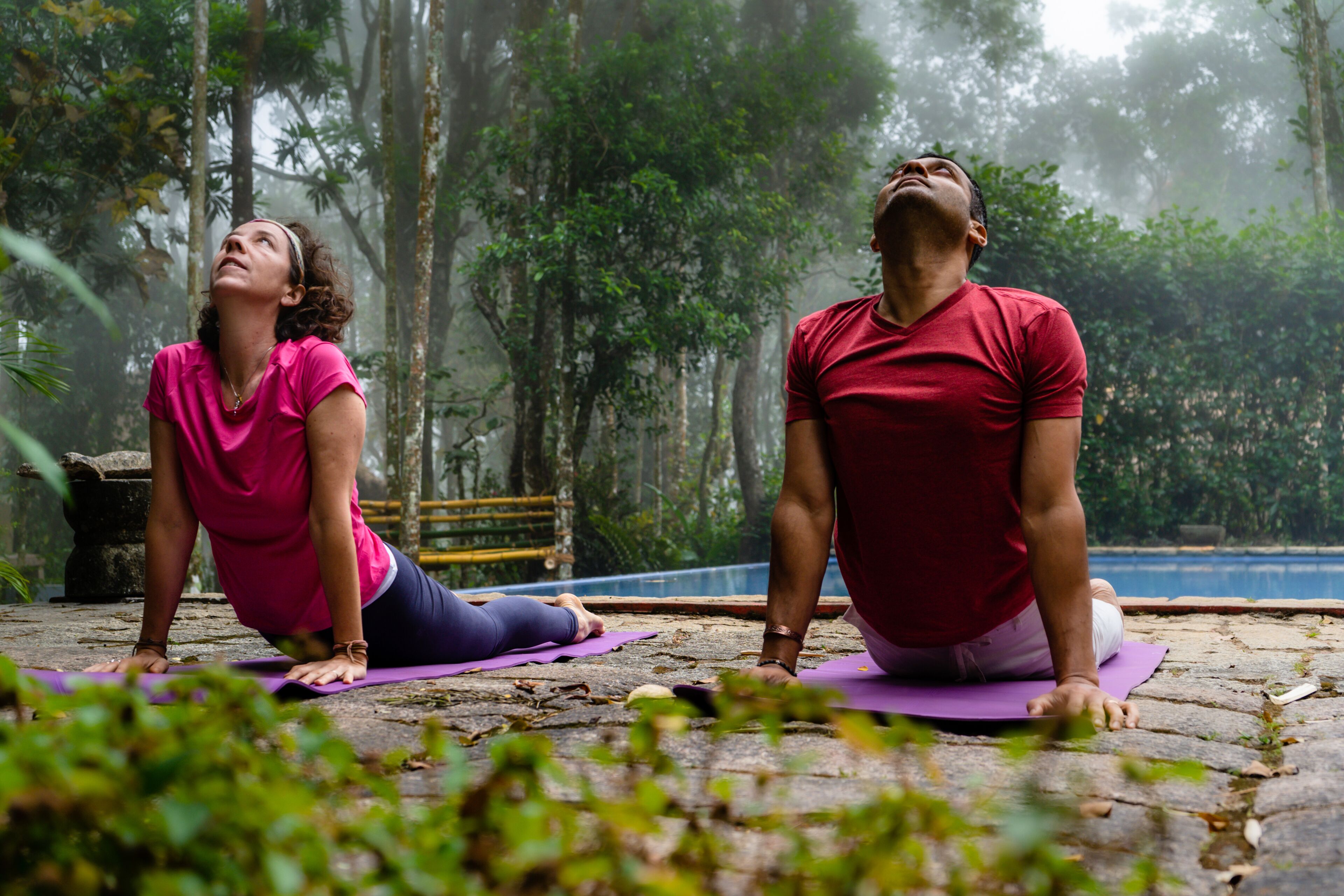 Dos personas sobre esterillas de yoga en un área boscosa con un sendero de piedra y un banco al fondo.
