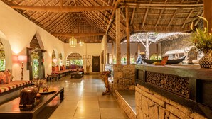 Um interior tradicional com telhado de palha, paredes de pedra e uma área de estar com almofadas e uma mesa de madeira.