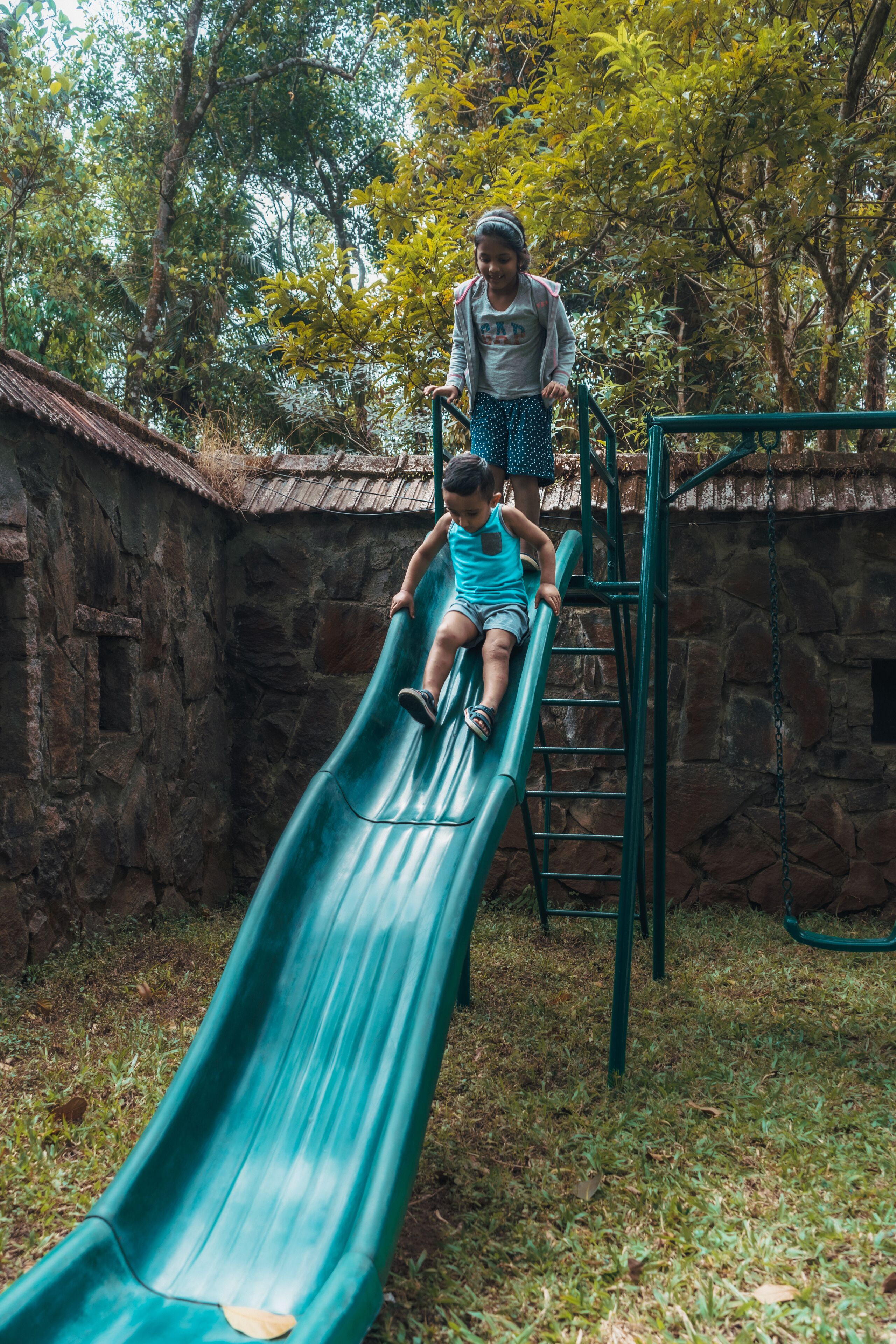 Un enfant descend en glissant sur un toboggan vert dans un parc, avec des murs en pierre et des arbres en arrière-plan.