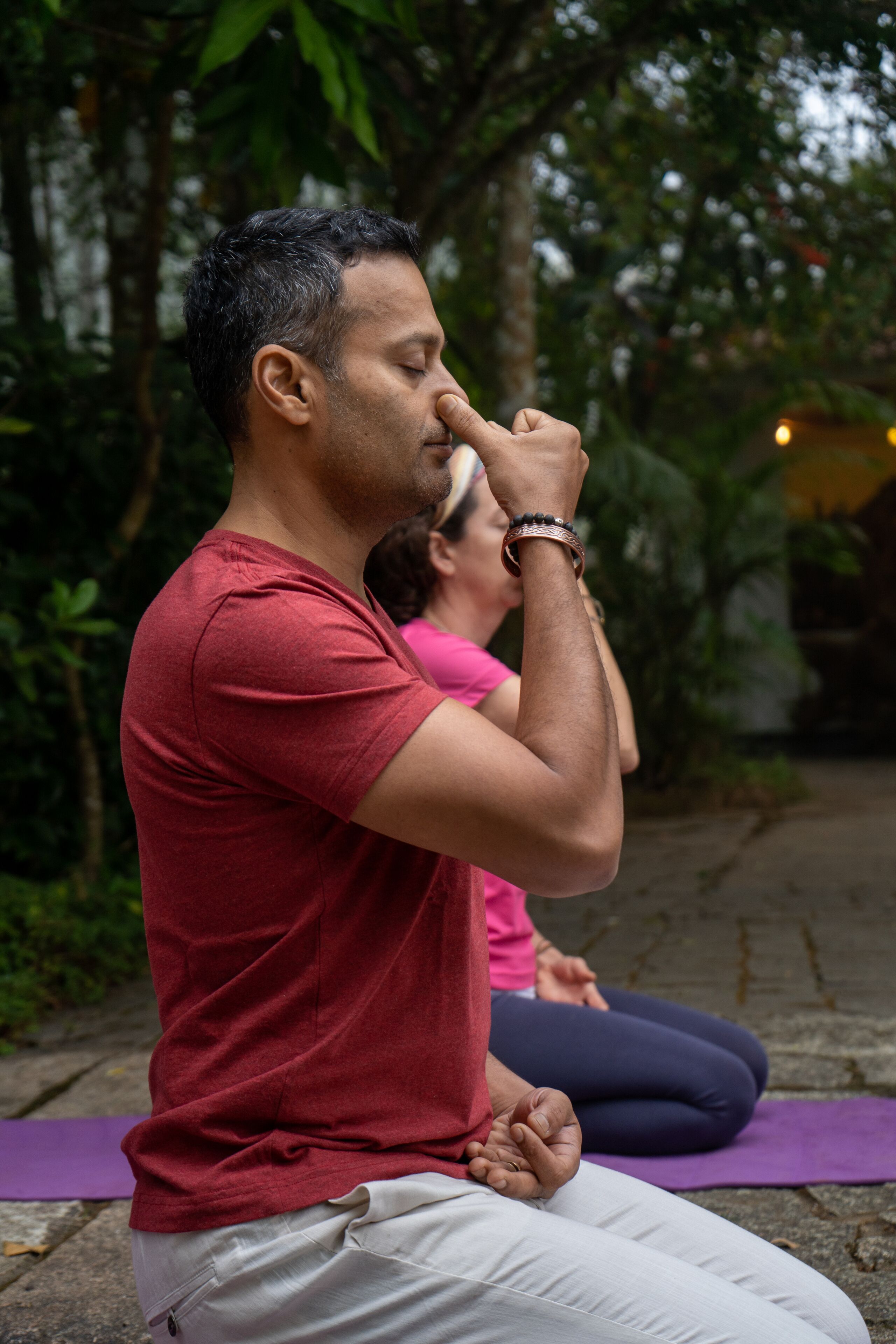 Dos personas sentadas sobre esterillas de yoga al aire libre, una con una camiseta roja y la otra con una rosa, con follaje verde de fondo.