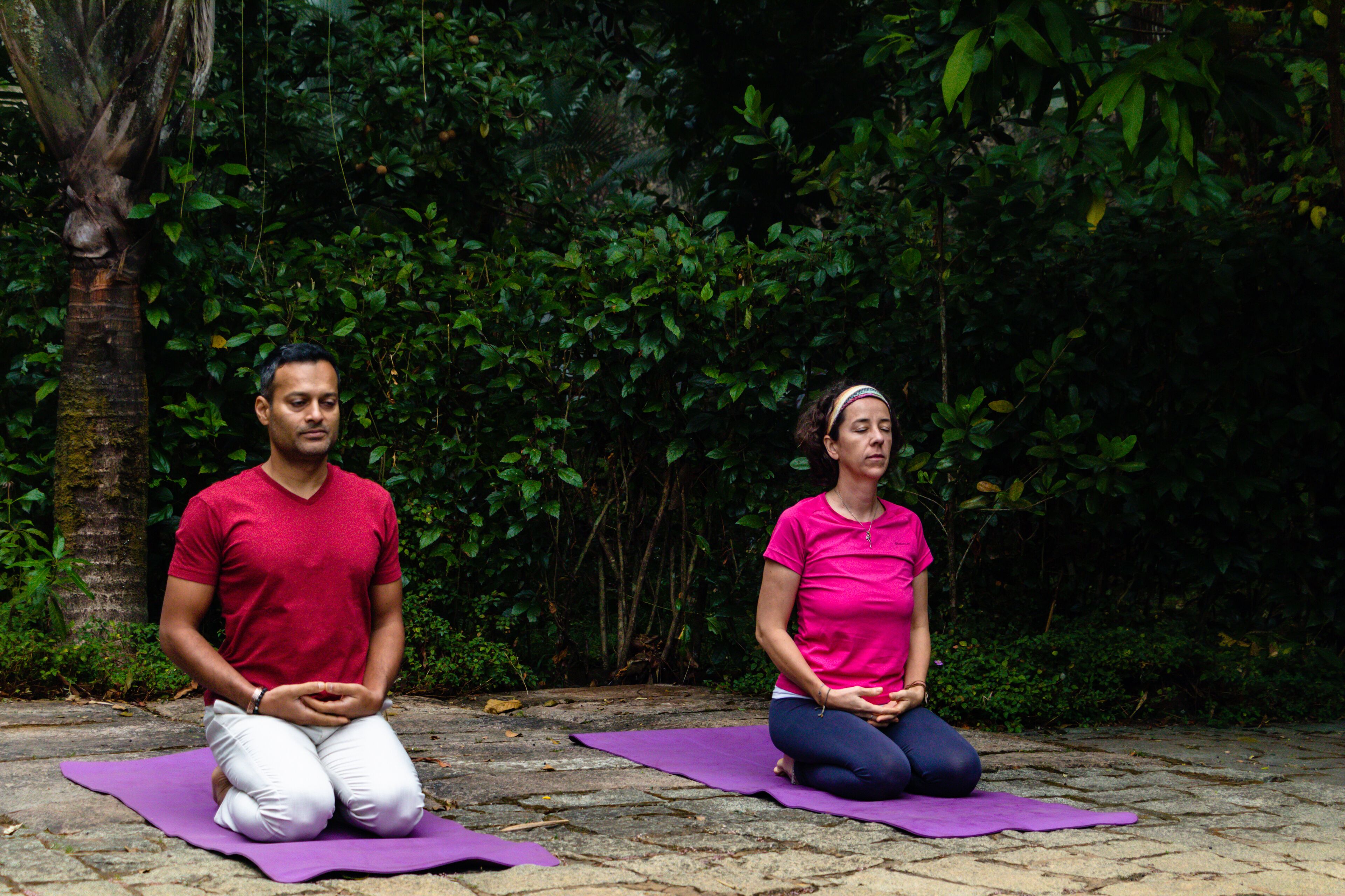 Two people sitting on yoga mats outdoors with dense foliage in the background.