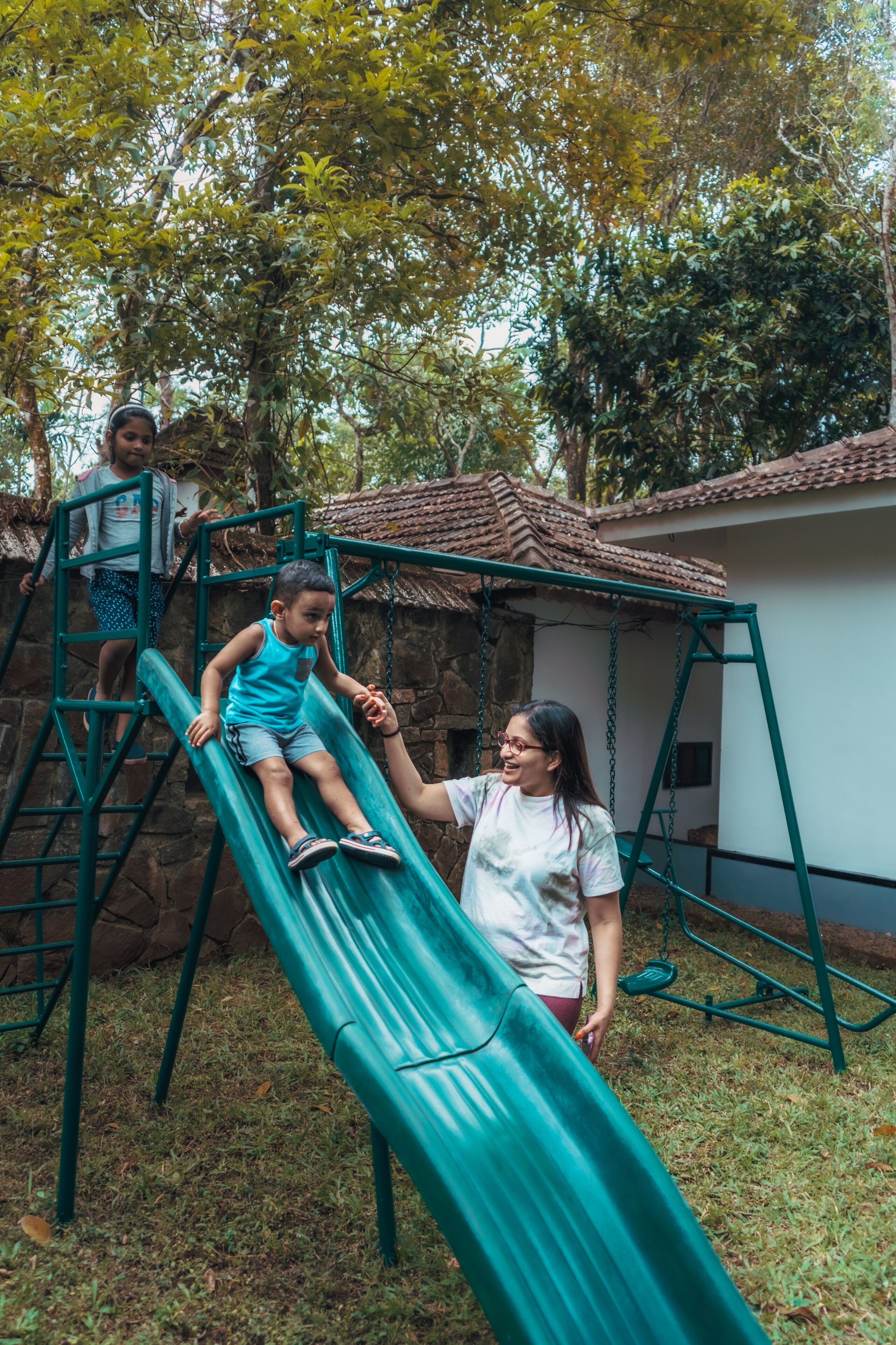 A playground with a green slide, a climbing structure, and a building with a tiled roof in the background.