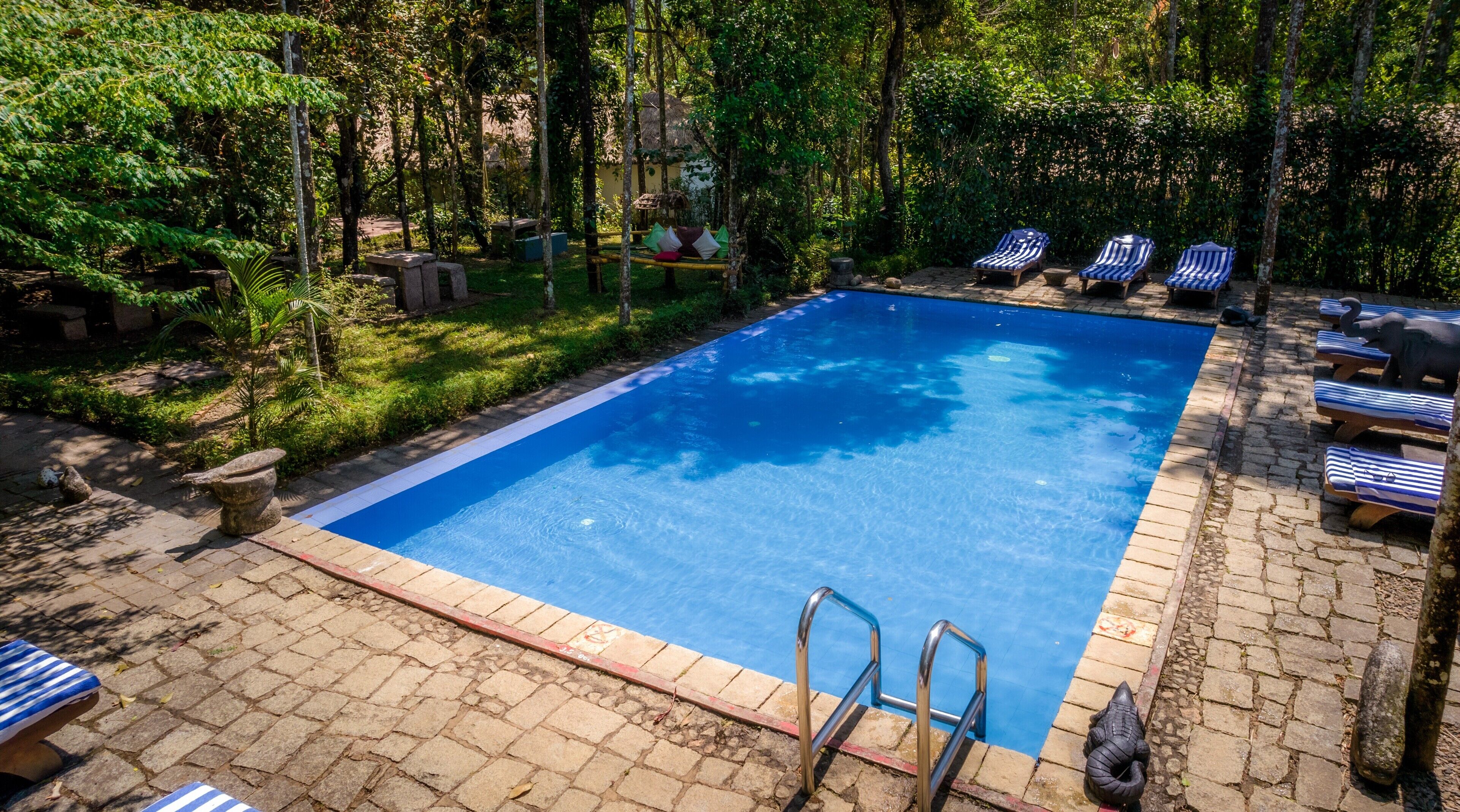 Une piscine rectangulaire aux parois bleues, entourée d’une terrasse en briques. On y trouve des chaises longues et une petite construction en pierre à proximité de la piscine.