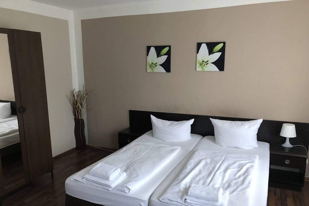 A hotel room with two single beds, a nightstand with a lamp, a vase with dried plants, and two wall-mounted flower paintings.