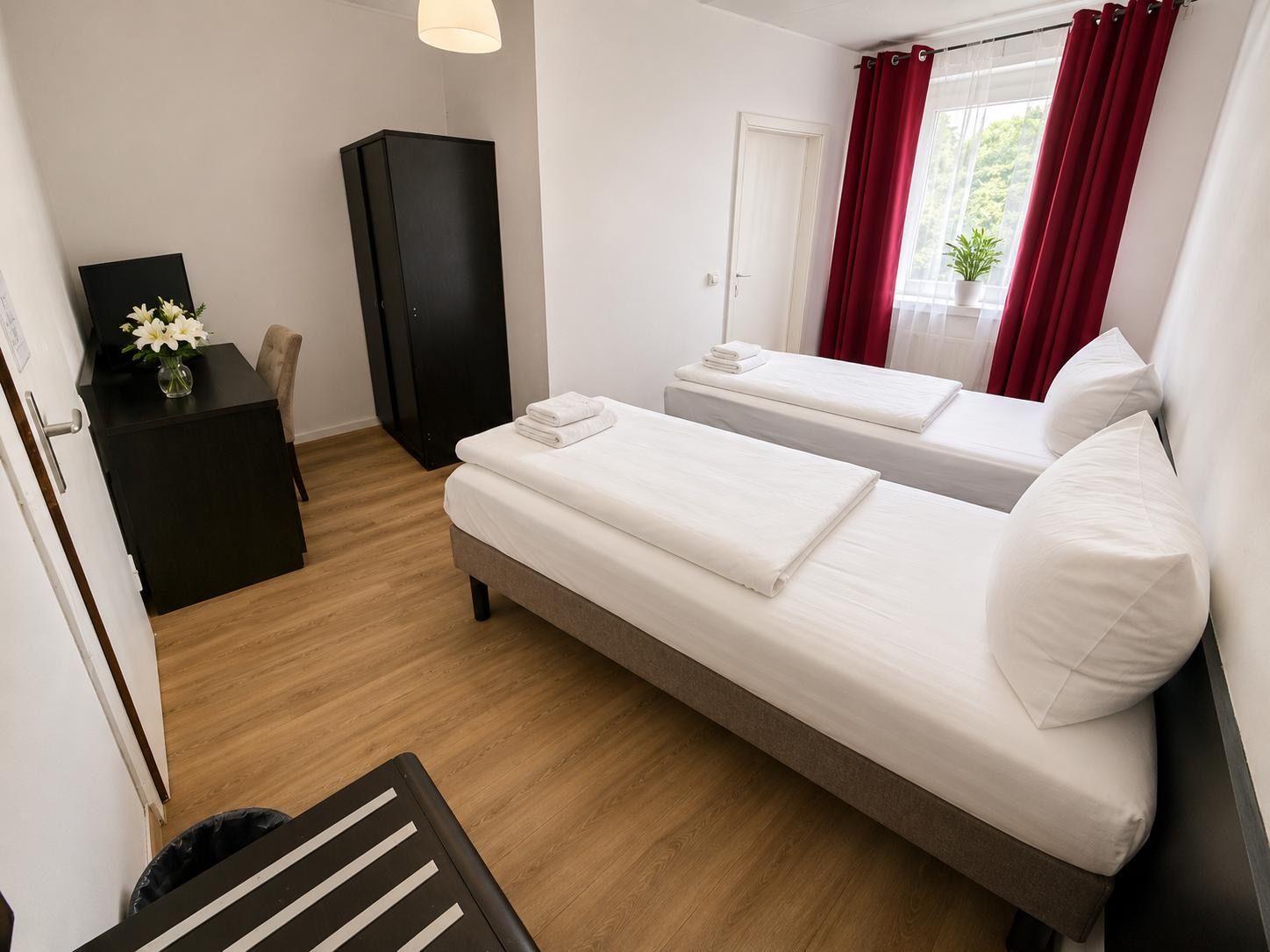 Ein Hotelzimmer mit zwei Betten, einem Kleiderschrank, einem Schreibtisch mit Blumenvase und Blick auf Grünfläche.