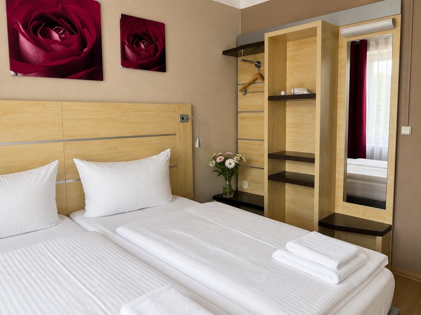 A hotel room with a bed, two pillows, a nightstand, a vase with flowers, and a wardrobe with a mirror.