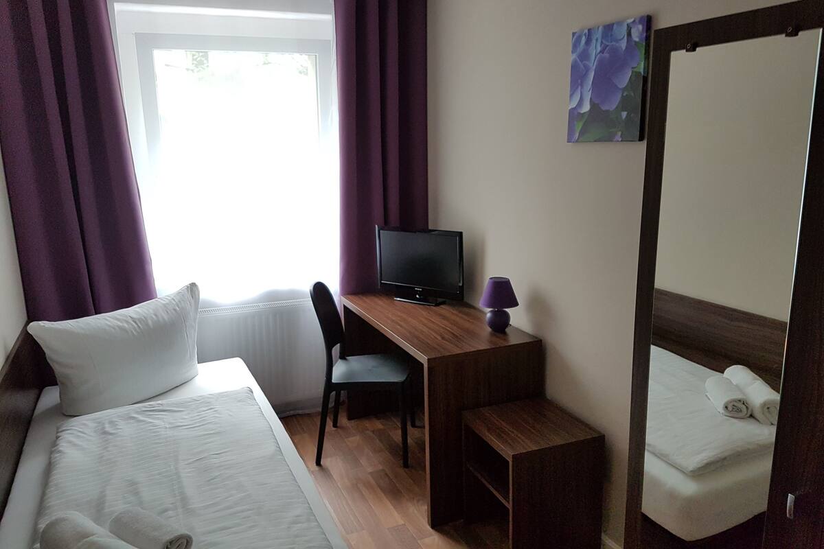 A hotel room with a bed, a desk with a computer, a chair, a small table, and a mirror.