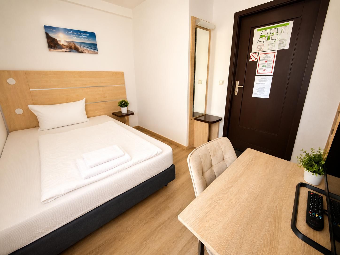 Ein Hotelzimmer mit einem Bett, einem Schreibtisch, einem Stuhl, einer kleinen Pflanze und einer Tür mit Aushängen.