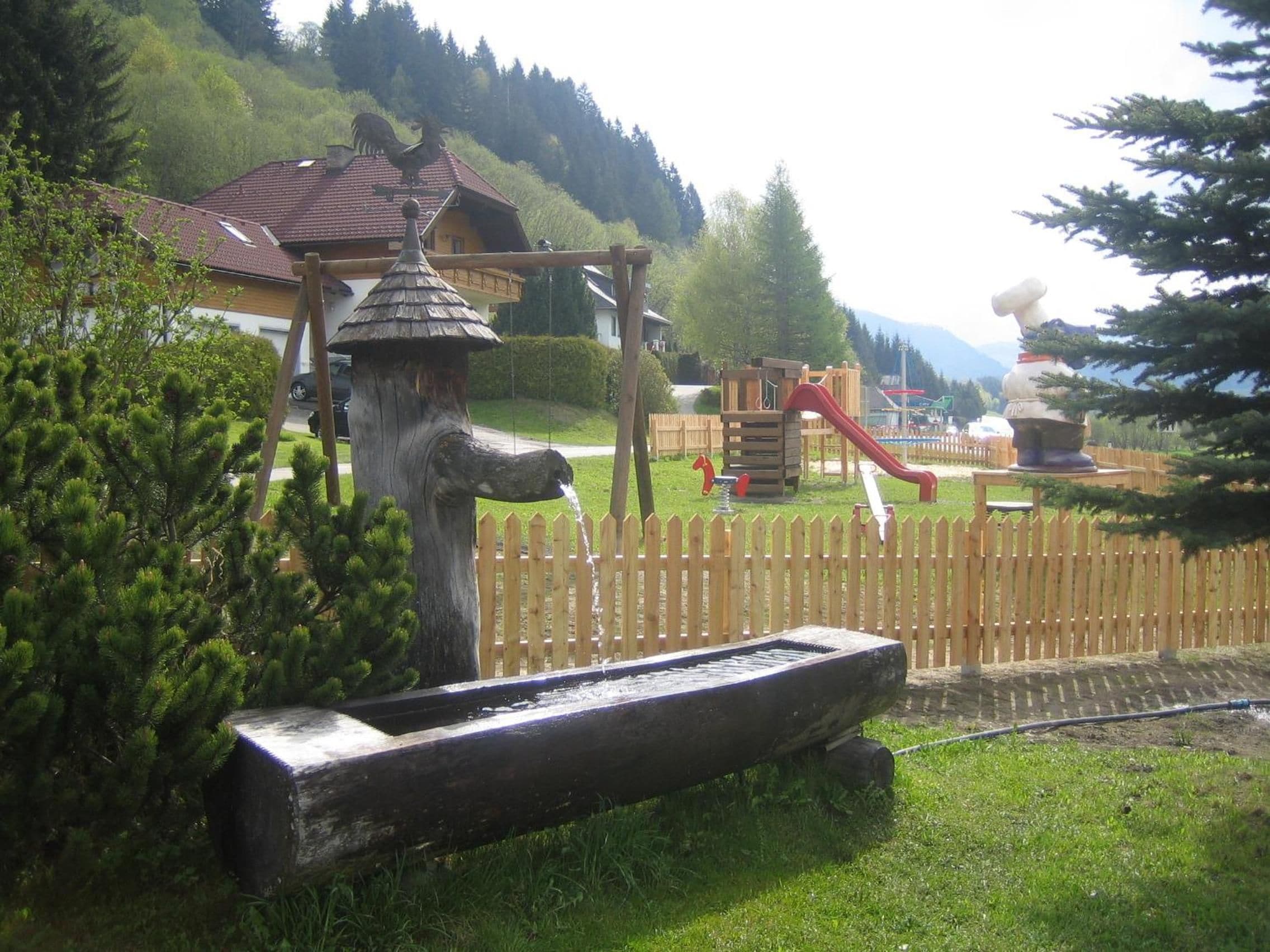 A wooden play structure with a slide and climbing elements, surrounded by a wooden fence and greenery.