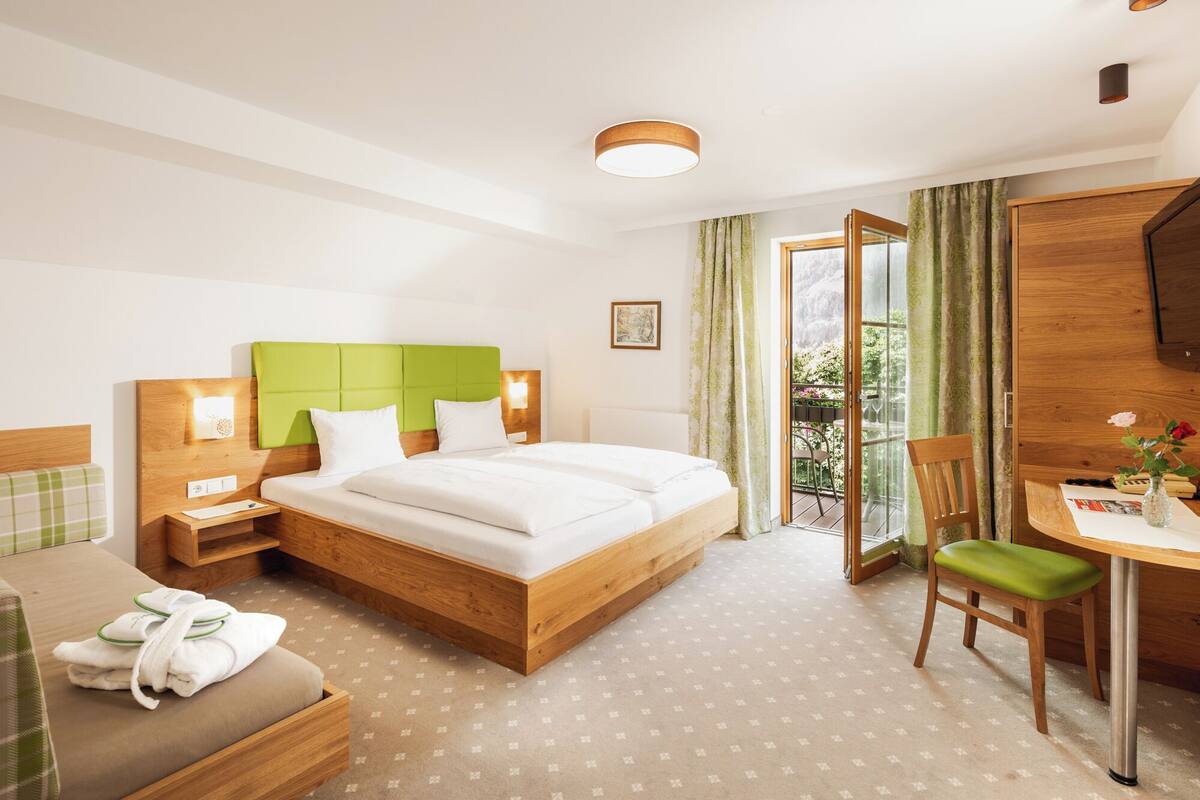 Habitación de hotel con una cama y un escritorio de madera, una silla, una mesita con un florero, y vista al exterior a través de una puerta de cristal.